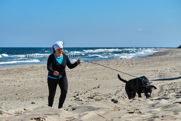 Kobieta spaceruje po plaży w wietrzny dzień z dużym czarnym psem.