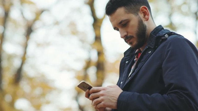 adult attractive man male using smart phone side view outside blurred trees park background typing texting message tapping fingers touchscreen information technology freelancer walking leisure app