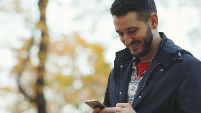 Portrait young smiling satisfied business man using mobile smart phone outdoors blur background nature happy cheerful bearded guy chatting online typing message sms communicate friends touching screen