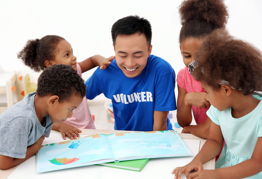 Young Male Volunteer Reading Book With Little Children In Light Room. Volunteering Abroad Concept