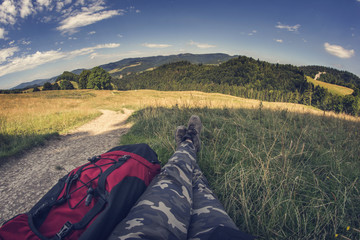 Trip to the mountains. Woman sitting on a grass with red backpack looking at the beautiful landscape. Legs with moro trousers