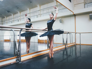 Ballerina in black tutu and pointe stretches on barre in ballet gym. Woman standing near bar and mirror, preparing for perfomance.