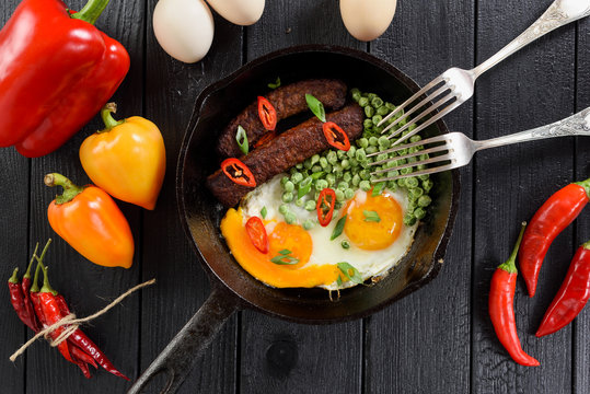 Nutritious protein meal. Fried eggs, peas, sausage prepared in cast iron pan served with bright bell and chili peppers on black background