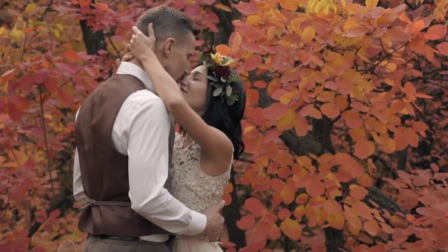 A man in a suit gently kisses a girl in a white dress and wreath of flowers standing in the middle of an autumn park, slow motion