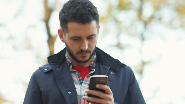 Young man standing park autumn using modern digital smart phone mobile serious calm expression face portrait checking news notification application gadget technology wifi 3g os pda downloading program