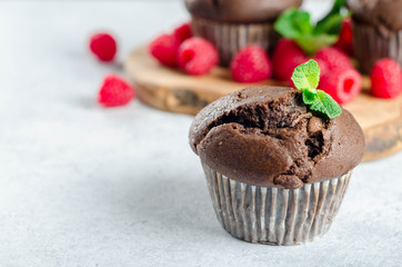 One chocolate muffin on a foreground and two muffins on a wooden board with fresh raspberries on the background, copy space for your text