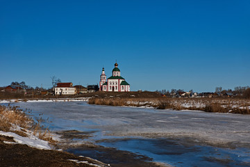 Orthodox temple on the river bank/City landscape of Suzdal. River, ice-bound,In the flood plain lies snow. In the distance, on a hill near the river stands Orthodox church.Suzdal.Golden Ring of Russia