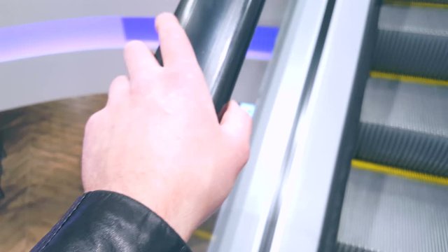 Male hand in a leather jacket keep the handrail of a traveling escalator in a modern bright spot. Blurred background