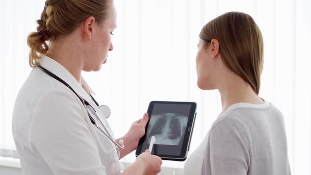 Woman physician with stethoscope talking with female patient in clinic. Female professional doctor at work. Showing X-ray on tablet. Health care
