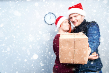 Spirit of Christmas and new year. Concept of a holiday and days off. A loving couple in Santa Claus hats. Guy hugging girl and holding gift in hand, focus on gift. with snowfall