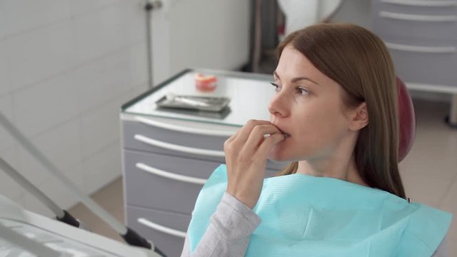 Woman patient in dental clinic. Waiting in dental chair for professional doctor stomatologist. Anxious, afraid, biting her nails. Dental equipment on background. Dental check up