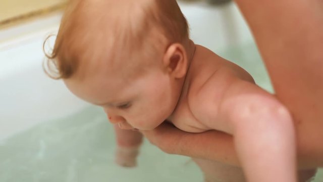 Cute baby boy taking bath mother hands pouring water back child half year old infant kid health care chubby cheeks beautiful big blue eyes looking camera closeup enjoying bathing wash clean skin body