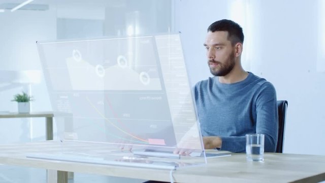In the Near Future Man Sitting at His Workstation, Works on His Modern Computer With Transparent Display. Display Shows User Interface with Interactive Charts and Graphics. 