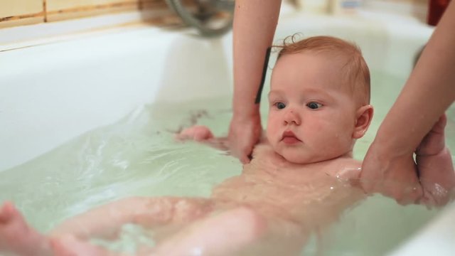nice adorable baby boy sliding back water mother hands support child taking bath tub bathing home calm infant intersted look pretty face chubby cheeks healthy hygiene measures gentle skin washed child