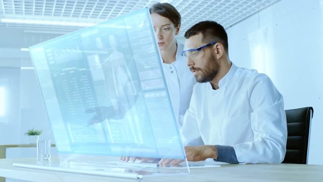 In the Futuristic Laboratory Male and Female Scientists Work on Transparent Computer Display, They Try to Prolong Human Life. Screen Shows Various Human Related Infographics and Data. 4K UHD.