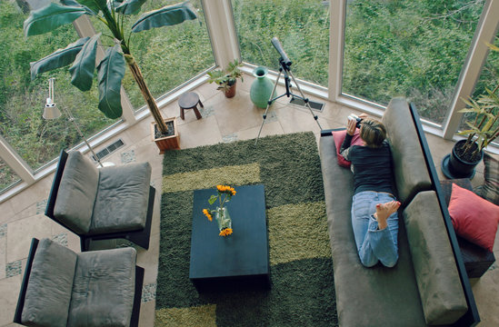 Woman laying on the couch at home looking through binoculars out the windows