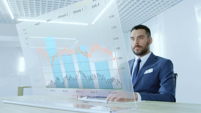 In the Near Future Successful Businessman Works on Computer with Transparent Display that Shows Interactive Line Charts and Other Useful Information. His Office is Bright and Modern. 