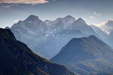 Fototapeta na wymiar Triglav, highest peak of Slovenia, from the north with snowy Rjavina and Cmir peaks, green forests of Jerebikovec and deep Kot Valley in afternoon sun light, Triglav National Park, Julian Alps, Europe