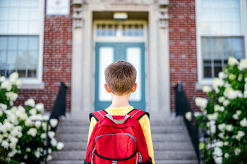 Boy wearing red backpack stands looking at the front entrance of a school