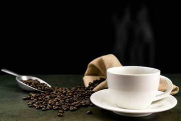 Hot coffee in the white cup with roast coffee beans, bag and scoop on stone table in black background
