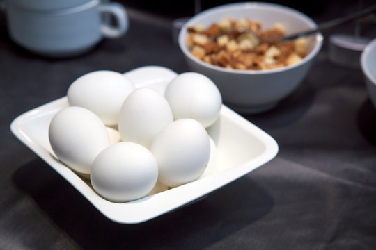 Close-up on a white plate lay boiled white eggs on a brown breakfast table, in the background a bowl with rusks