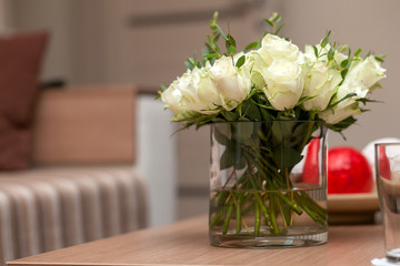 Close-up of a beautiful bouquet of fresh white roses on short stems in a transparent glass vase on a wooden table stand in the bedroom, in the background a bed