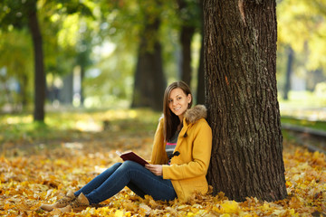 A beautiful happy smiling brown-haired woman in yellow coat and jeans sitting under the maple tree with a red book in fall city park on a warm day. Autumn golden leaves. Reading concept