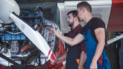 Two mechanics working on a small aircraft in a hangar with the cowling off the engine as they...