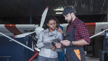 Worker man and woman standing with tablet and talking in hangar.