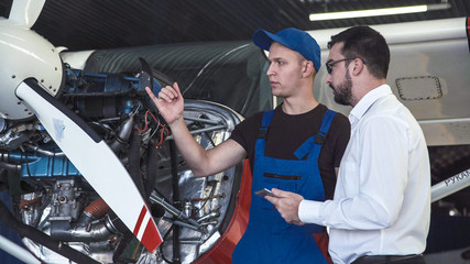 Mechanic standing near plane, looking at engine and talking to draftsman.