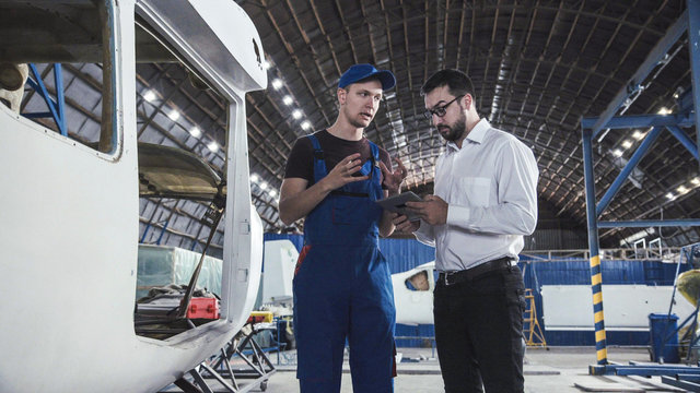 Aircraft mechanic and draftsman standing near plane carcass and talking with each other.