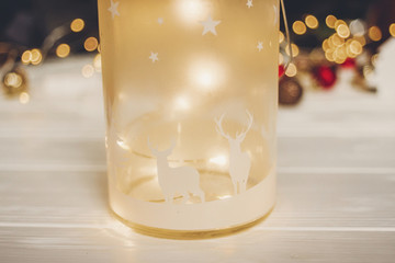 christmas present. lantern with deers and simple ornaments and christmas tree on white wood with lights in background, space for text. seasonal greetings. happy holidays. xmas gift.