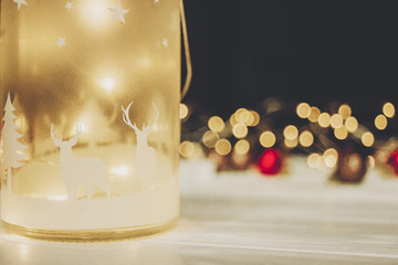 christmas atmospheric moment. lantern with deers and simple ornaments and christmas tree on white wood with lights in background, space for text. seasonal greetings. happy holidays