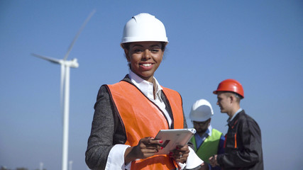 Female engineer wearing hardhat standing with digital tablet against other engineers and wind...