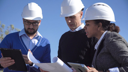 Group of engineers wearing helmets discussing over plans outdoors