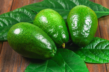 Three green raw ripe avocados with leaves lie on a wooden brown table.