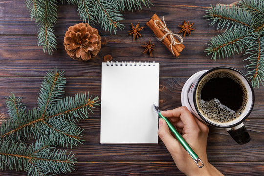 Woman write wishes or to do list in notebook, coffee mug, Christmas. christmas tree branches, pine cones, red berries, on marble table, copy space top view