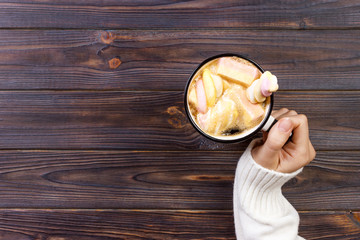 female hand holding cup of hot cocoa or chocolate with marshmallow on wooden table from above