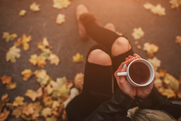 A young girl holding a cup of hot drink and smiling at the background of a autumn forest. Attractive young woman in autumn outdoor. Female model drinking hot tea in the autumn forest, close up