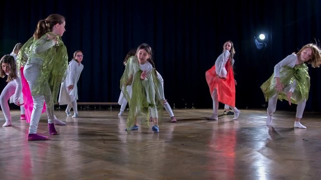 Girls are wearing white costumes and capes of different colours and are performing in a school play and are dancing hip hop and modern dancing.