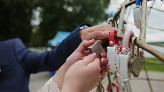 Groom hanging wedding padlock on the pipe near a variety of padlocks. Wedding colorful locks hanging on the pipes of a pedestrian bridge the background a river. Close-up