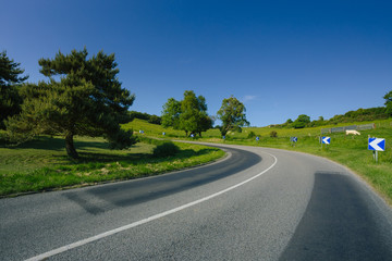 Fototapeta na wymiar Empty asphalt curvy road passing through green fields and forests. Countryside landscape on a sunny spring day in France. Sunbeams in the sky. Transport, industrial agriculture, road network concept