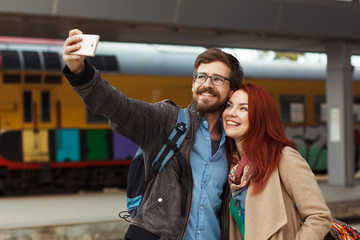 Young couple hiking taking selfie with smart phone. Happy young man and woman taking self portrait with travel station in background.