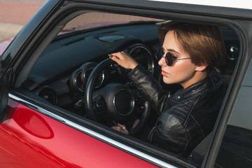 Fototapeta na wymiar Beautiful girl in leather jacket and sun glasses is looking away while sitting in her car