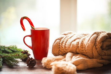 Christmas Hot Coffee in red cup on wooden table on a frosty winter day window background with knitted scarf, candy  and christmas tree branches. Home cozy holidays background.