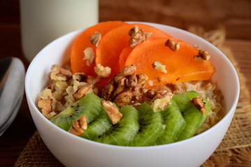 Oatmea porridge with toppings. Oatmeal with fresh persimmon, kiwi and walnuts in a bowl. A bottle of milk, a spoon on a wooden table. Healthy porridge for Breakfast. Closeup