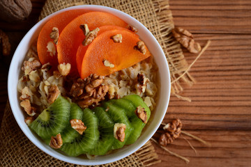 Oatmeal with fruit. Oatmeal with sliced persimmon, kiwi and walnuts in a bowl and on a wooden table. Healthy cereal for weight loss. Top view