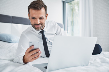Smiling businessman using phone while lying on bed in hotel room on business trip
