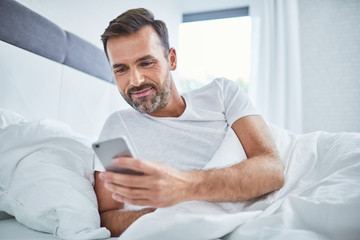 Handsome man using phone in bed after waking up in the morning