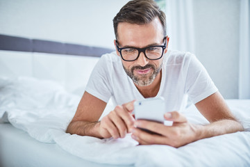 Handsome man using phone lying on bed in the morning